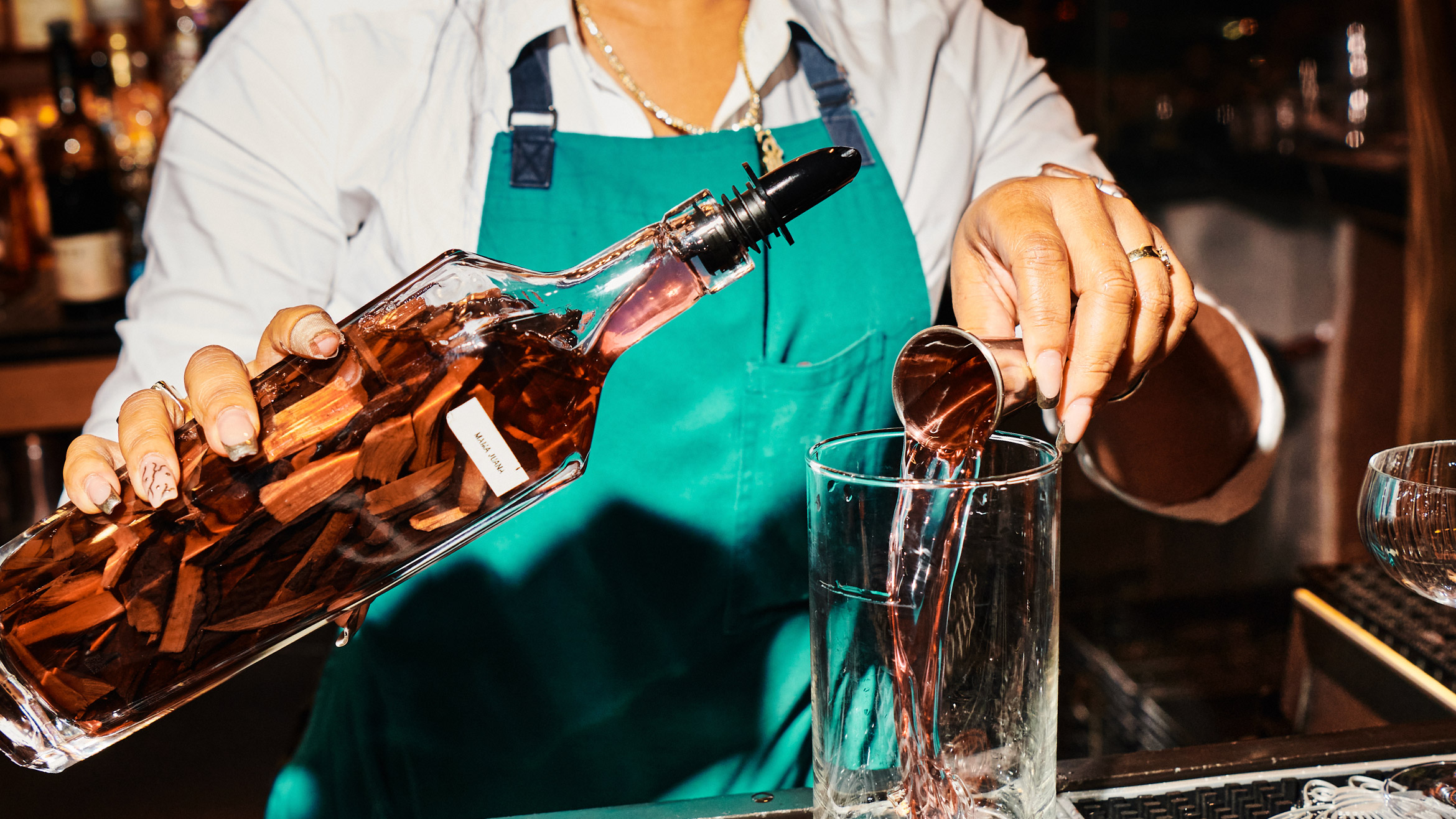 A bartender pours a jigger of liquid into a glass, in the other hand they hold a bottle filled with a brown liquor and botanicals.
