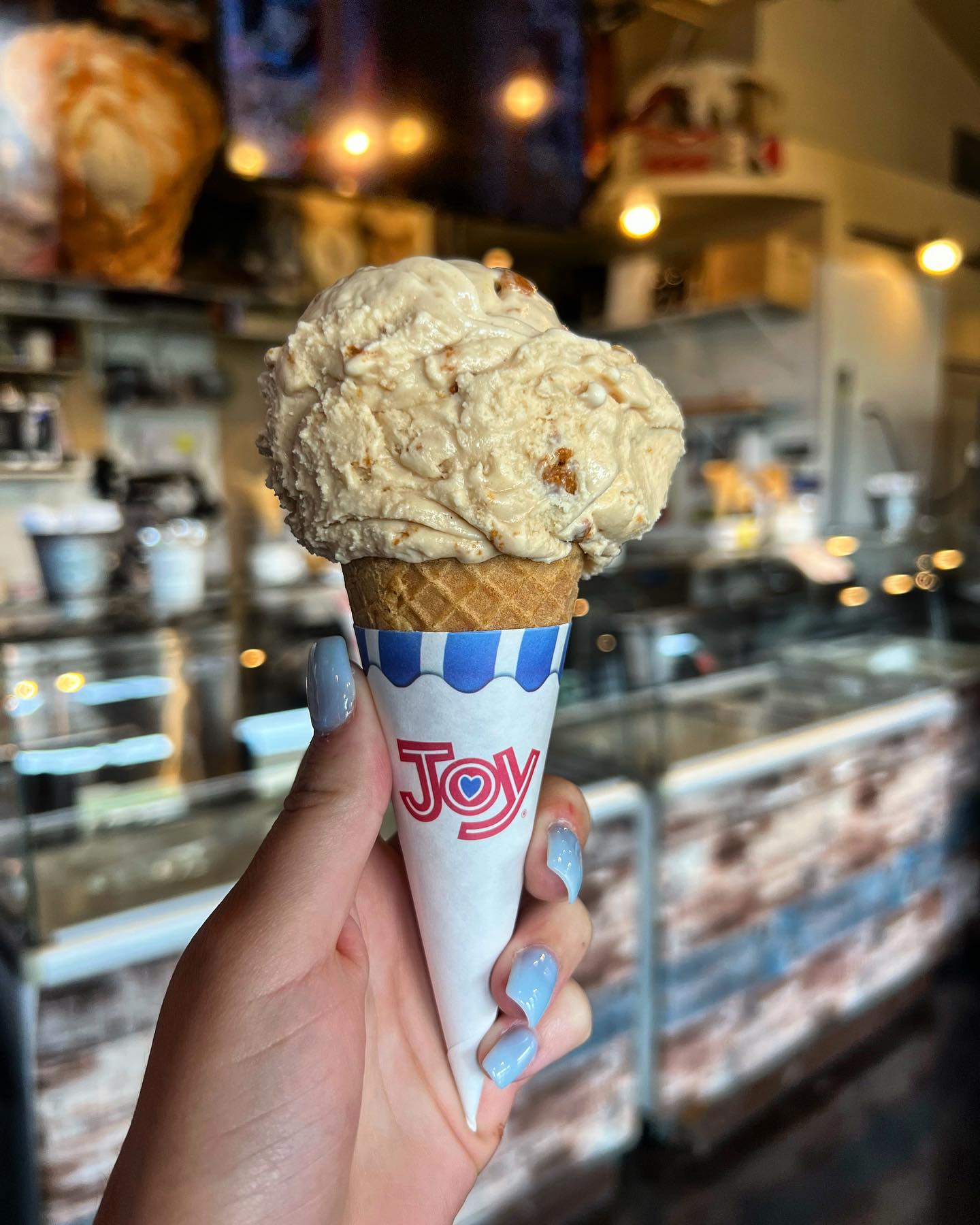 An ice cream (looks like butter pecan) in a cone held up with an ice cream shop in the background.