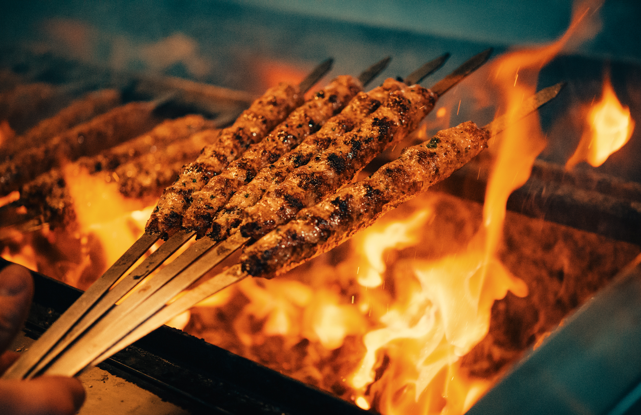 A hand holds skewers of meat over a ranging fire grill.
