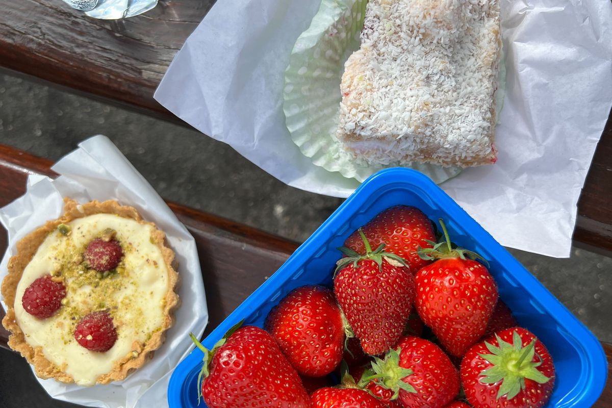 A picnic spread shows an array of coconut-dusted cookies, ultra-ripe strawberries, and a cream tart studded with three raspberries spread out on a picnic blanket.
